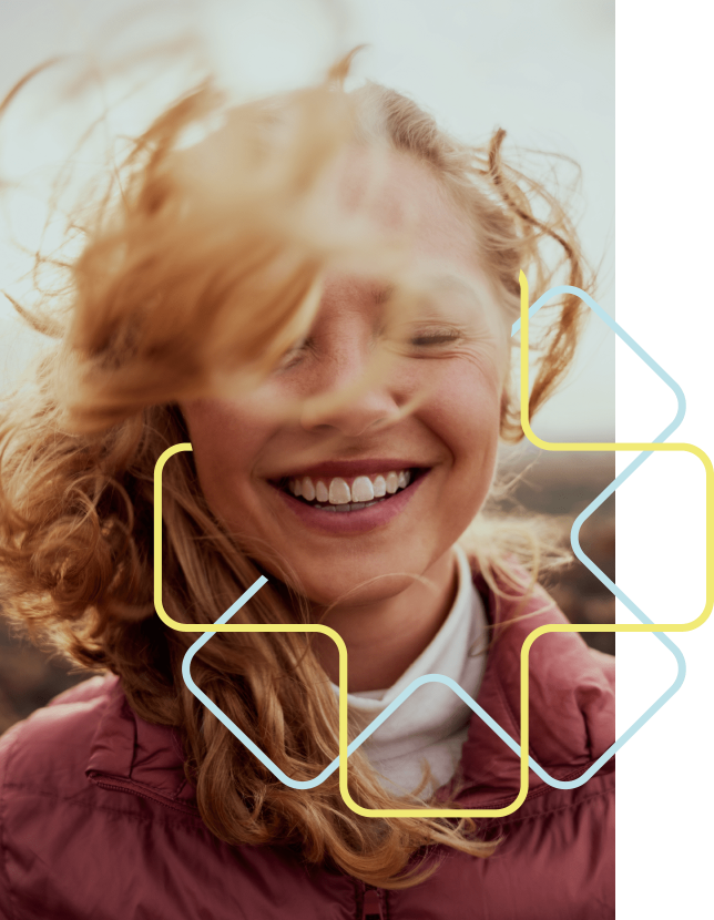 Das Gesicht einer lächelnden jungen Frau mit geschlossenen Augen, der Wind weht ihr rotes Haar ins Gesicht. Sie trägt eine Daunenjacke in altrosa. Der Hintergrund ist verschwommen. Das geometrische Stilelement in gelb und hellblau umrahmt ihr Gesicht..