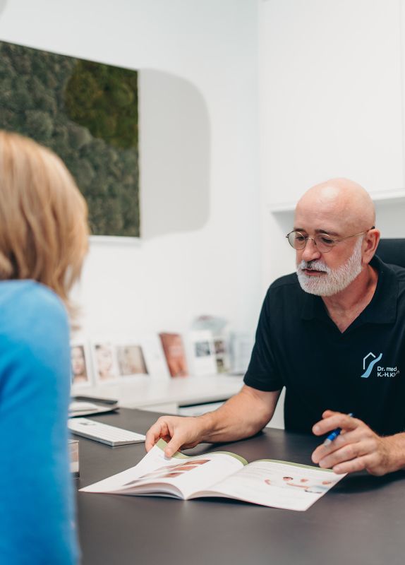 Foto von Dr. Klein, der am Schreibtisch sitzt und in einer Broschüre blättert. Im Gegenüber sitzt eine Patientin mit blauen Oberteil. Der Hintergrund ist verschwommen.