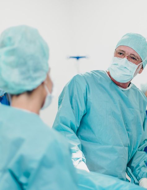 Herr Dr. Klein, in voller hellblauer OP-Montur mit Haube, Kittel und Maske blickt zu seiner Kollegin im Vordergrund, die ebenfalls OP-Kleidung trägt.