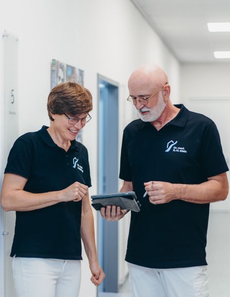 Frau Dr. Nölling und Herr Dr. Klein, stehen nebeneinander im Flur der Praxis und blicken gemeinsam auf ein Tablet