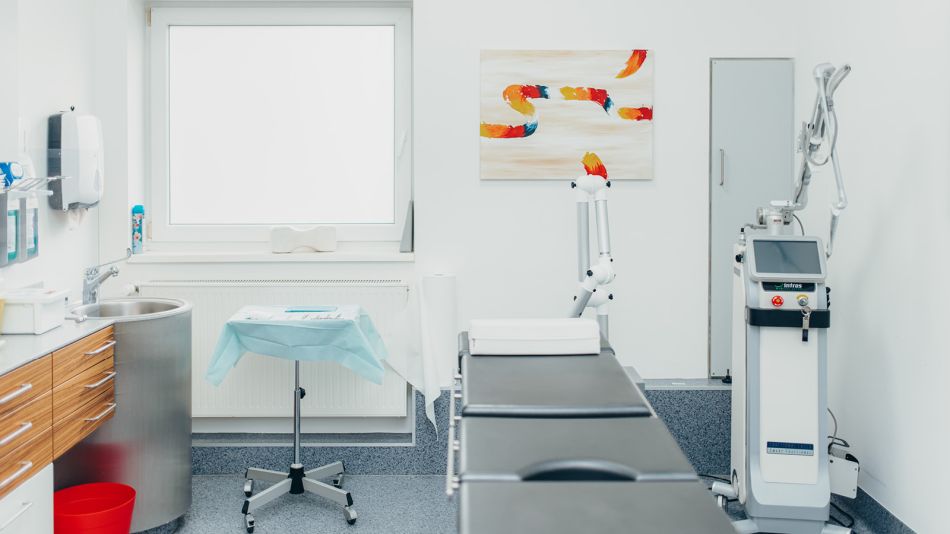 Ein Behandlungszimmer mit einer Liege in der Mitte des Raums, links steht ein medizinisches Gerät, rechts ein Tisch mit Instrumenten. An der Wand hängt ein buntes, abstraktes Gemälde. Ganz links ist ein Waschbecken zu sehen.