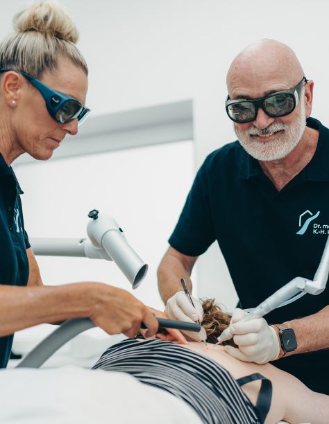 Herr Dr. Klein (rechts) und eine weitere Mitarbeiterin (links) behandeln den Rücken einer Patientin, der professionell mit medizinischen Geräten eine Warze entfernt wird. Beide tragen eine Schutzbrille, Dr. Klein lächelt in die Kamera.