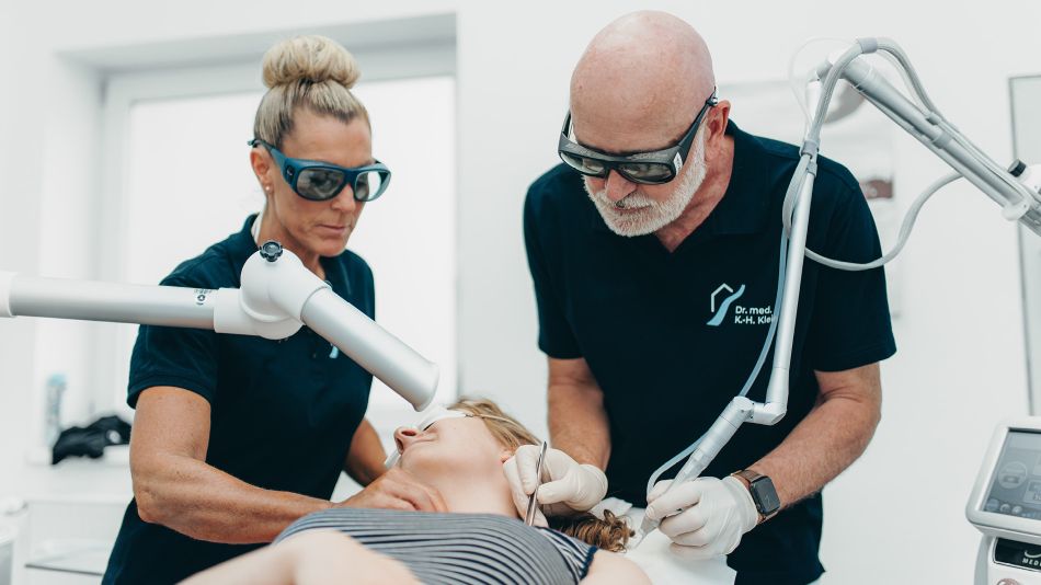 Herr Dr. Klein (rechts) und eine weitere Mitarbeiterin (links) behandeln eine Patientin an der Schulter. Beide tragen eine Schutzbrille und Handschuhe. Medizinischen Behandlungsgeräte sind im Vorder- und Hintergrund zu sehen.
