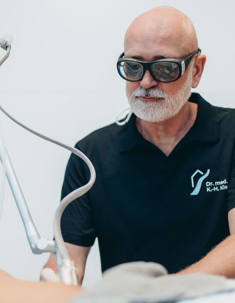 Herr Dr. klein trägt eine Schutzbrille und behandelt eine nicht sichtbare Person mit einem Lasergerät. Er trägt das dunkelblaue Shirt der Praxisklinik am Flüsschen.