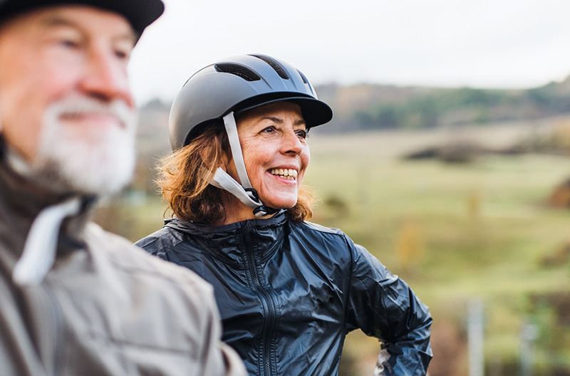 Ein älteres Paar ist auf Fahrradtour unterwegs. Der Mann, verschwommen im Vordergrund, hat einen grauen Bart, trägt einen Helm und eine dunkle Jacke. Die Frau im Hintergrund lächelt, trägt einen Helm und eine schwarze Jacke.