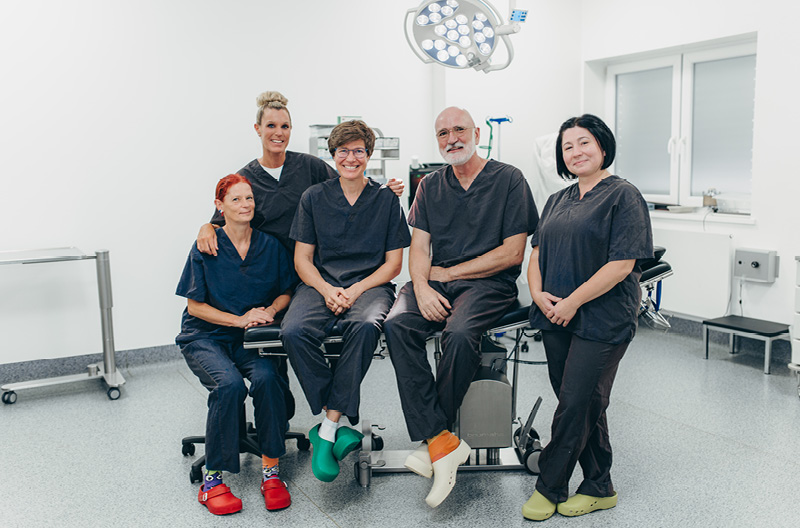 Ein Gruppenbild mit Dr. Klein und Dr. Nölling, die auf einer Behandlungsliege sitzen. Links neben Dr. Nölling sind zwei MFA, eine sitzt und die andere steht hinter den beiden. Rechts von Dr Klein steht eine MFA. Alle tragen dunkle OP-Kleidung.